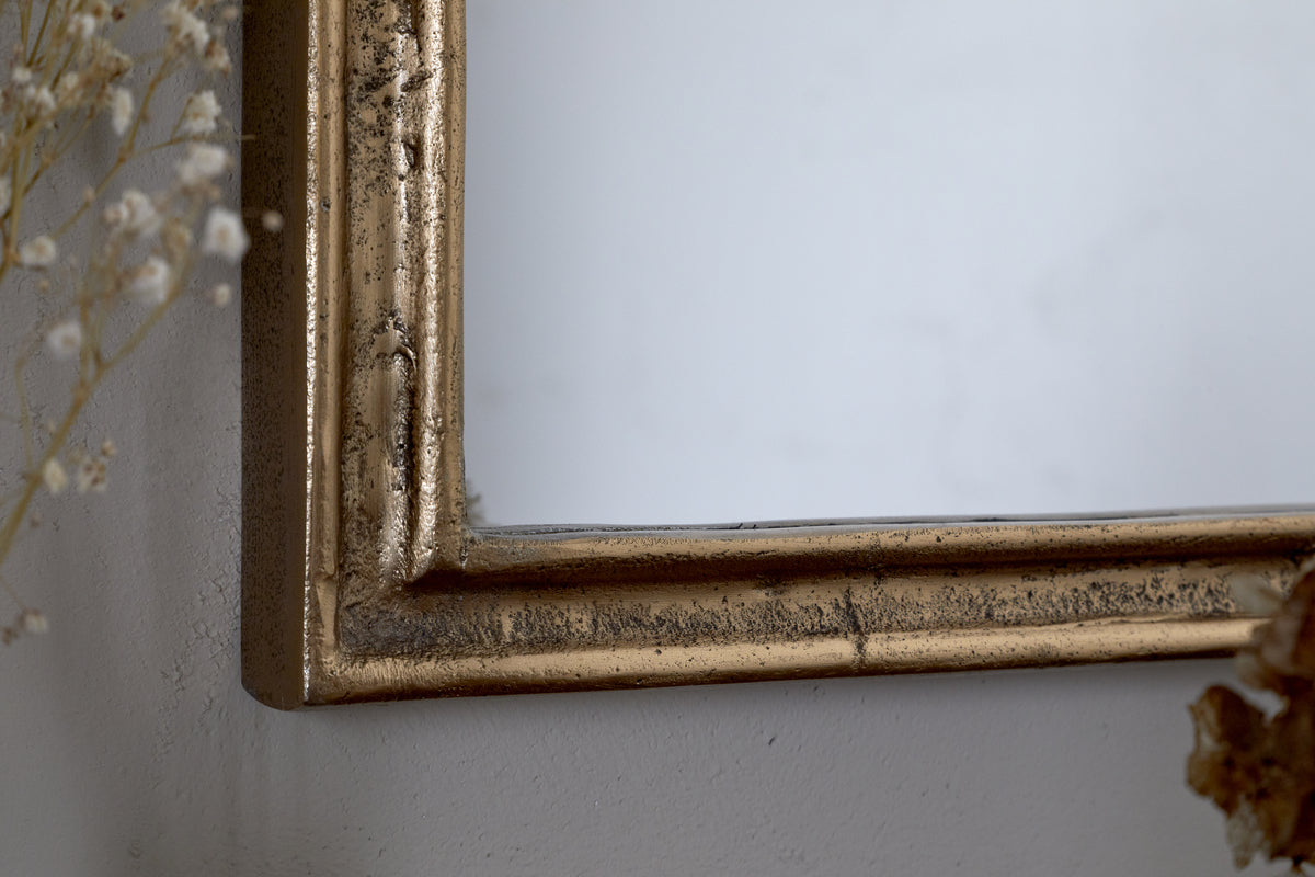 Close-up of the Almora Wall Mirror - Antique Brass (Extra Small) by nkuku, showing its slightly distressed frame against a light wall. Delicate dried flowers appear at the left and bottom edges.