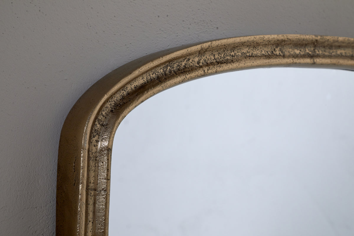 Close-up of the top corner of the nkuku Almora Wall Mirror, Extra Small, in antique brass with a distressed finish, set against a light gray wall.