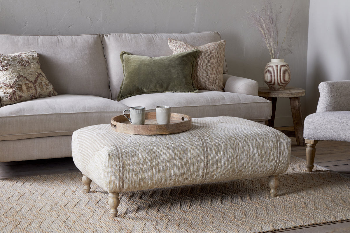 A cozy living room featuring a beige sofa, two accent pillows, the Aranya Upholstered Ottoman - Natural by nkuku, a round wooden tray with two mugs, textured rug, side table, and a ceramic vase with dried grasses for the perfect touch.