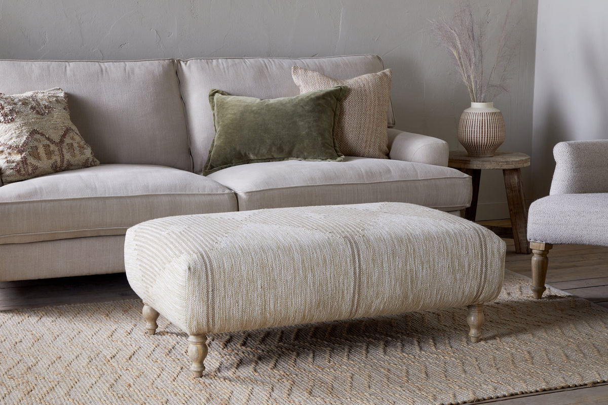 A cozy living room features a beige sofa with textured pillows, a matching armchair, and the nkuku Aranya Upholstered Ottoman - Natural as the perfect accent. A woven rug adds warmth, while a wooden side table displays a ceramic vase with dried grasses.
