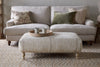 A beige sofa with three pillows sits against a light wall. In front is the nkuku Aranya Upholstered Ottoman - Natural, topped with a round wooden tray and two ceramic cups. The room features a woven rug and wooden accents.