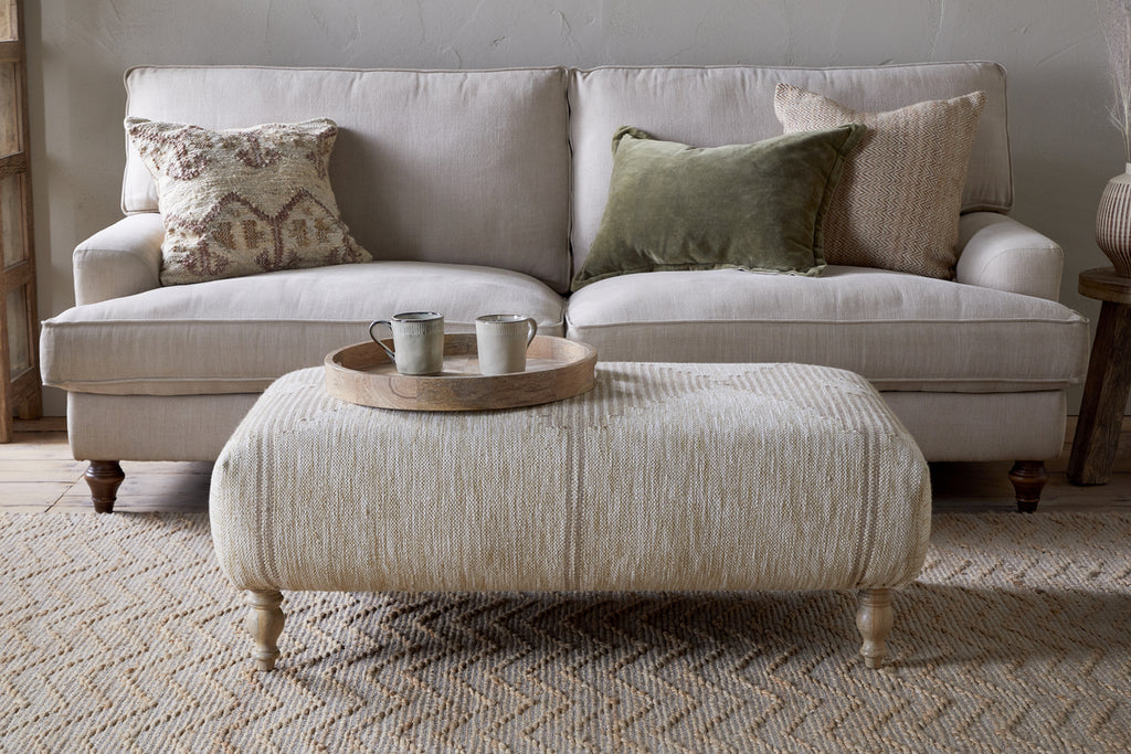 A beige sofa with three pillows sits against a light wall. In front is the nkuku Aranya Upholstered Ottoman - Natural, topped with a round wooden tray and two ceramic cups. The room features a woven rug and wooden accents.