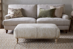 A neutral-toned living room features a light beige sofa with three textured pillows, paired with the Aranya Upholstered Ottoman – Natural by nkuku on a woven rug—a perfect living room accent.