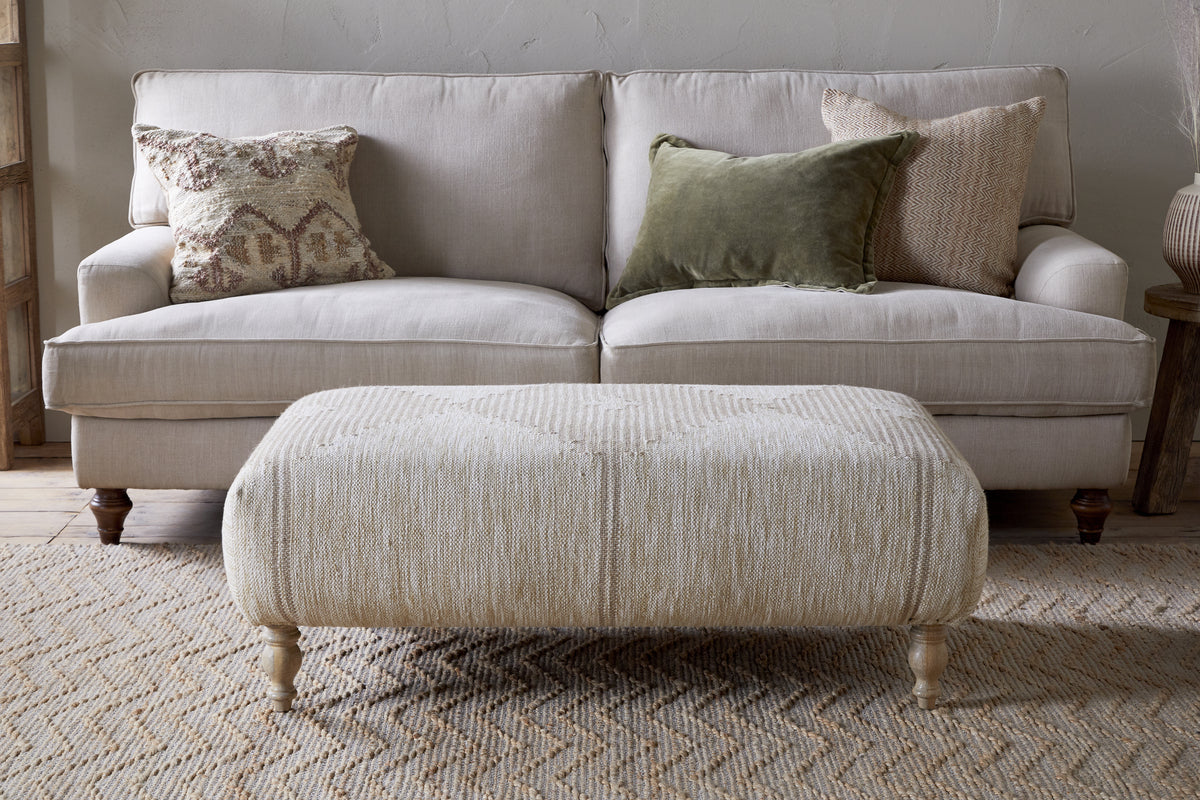 A neutral-toned living room features a light beige sofa with three textured pillows, paired with the Aranya Upholstered Ottoman – Natural by nkuku on a woven rug—a perfect living room accent.