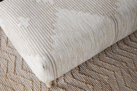 A close-up of the nkuku Aranya Upholstered Ottoman in Natural, featuring a subtle geometric pattern, sits on a tan and brown zigzag rug—making it an ideal stylish accent for your living room.