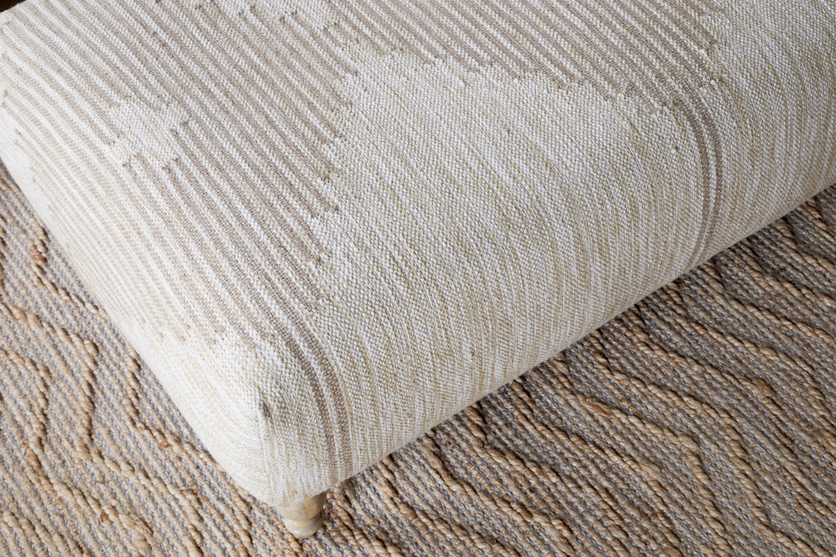 A close-up of the nkuku Aranya Upholstered Ottoman in Natural, featuring a subtle geometric pattern, sits on a tan and brown zigzag rug—making it an ideal stylish accent for your living room.
