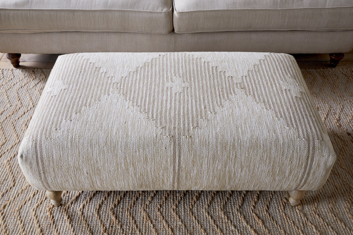 The Aranya Upholstered Ottoman - Natural by nkuku adds style to your living room with its beige textured fabric, subtle diamond pattern, and short wooden legs, standing out as a chic accent in front of a light-colored sofa.