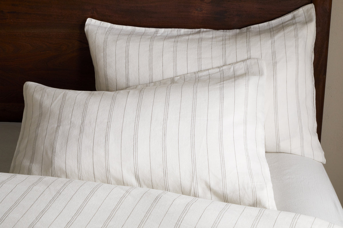 Two white pillows with thin, vertical gray stripes rest against a dark wooden headboard. Pair them with nkuku’s Anta Hemp & Cotton Housewife Pillowcases in Sage Green for a fresh touch. A matching striped blanket is partially visible below.