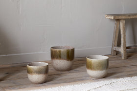 Three Almeida Reactive Glaze Plant Pots by nkuku in Olive & Natural, each a different size with an earthy brown-green to beige gradient glaze, are arranged on a wooden floor beside a rustic bench and a woven rug against a textured wall.