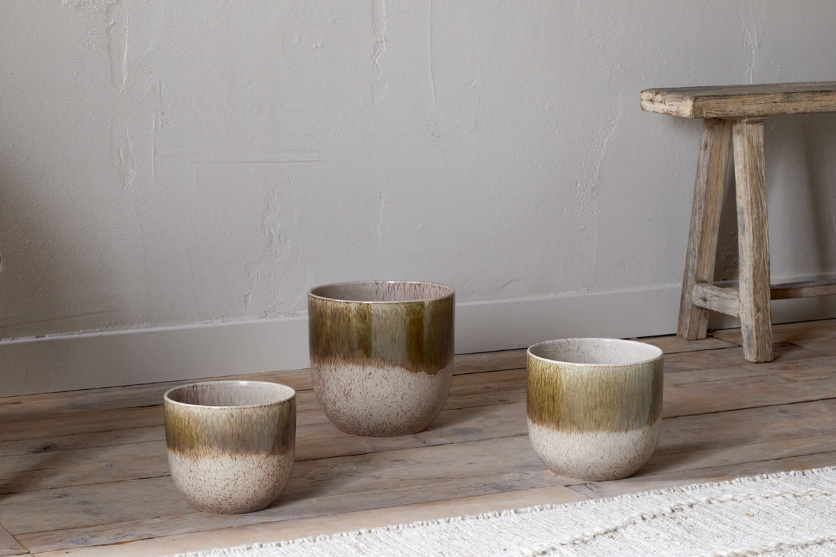 Three Almeida Reactive Glaze Plant Pots by nkuku in Olive & Natural, each a different size with an earthy brown-green to beige gradient glaze, are arranged on a wooden floor beside a rustic bench and a woven rug against a textured wall.