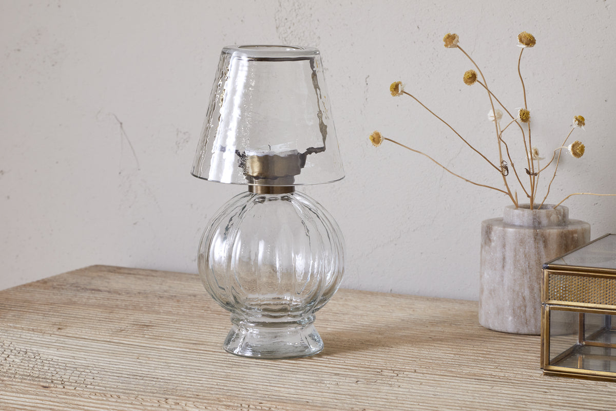 The nkuku Adesola Glass Tealight Holder Lamp in clear glass sits on a wooden surface beside dried yellow flowers and a glass box, set against a light, textured wall.
