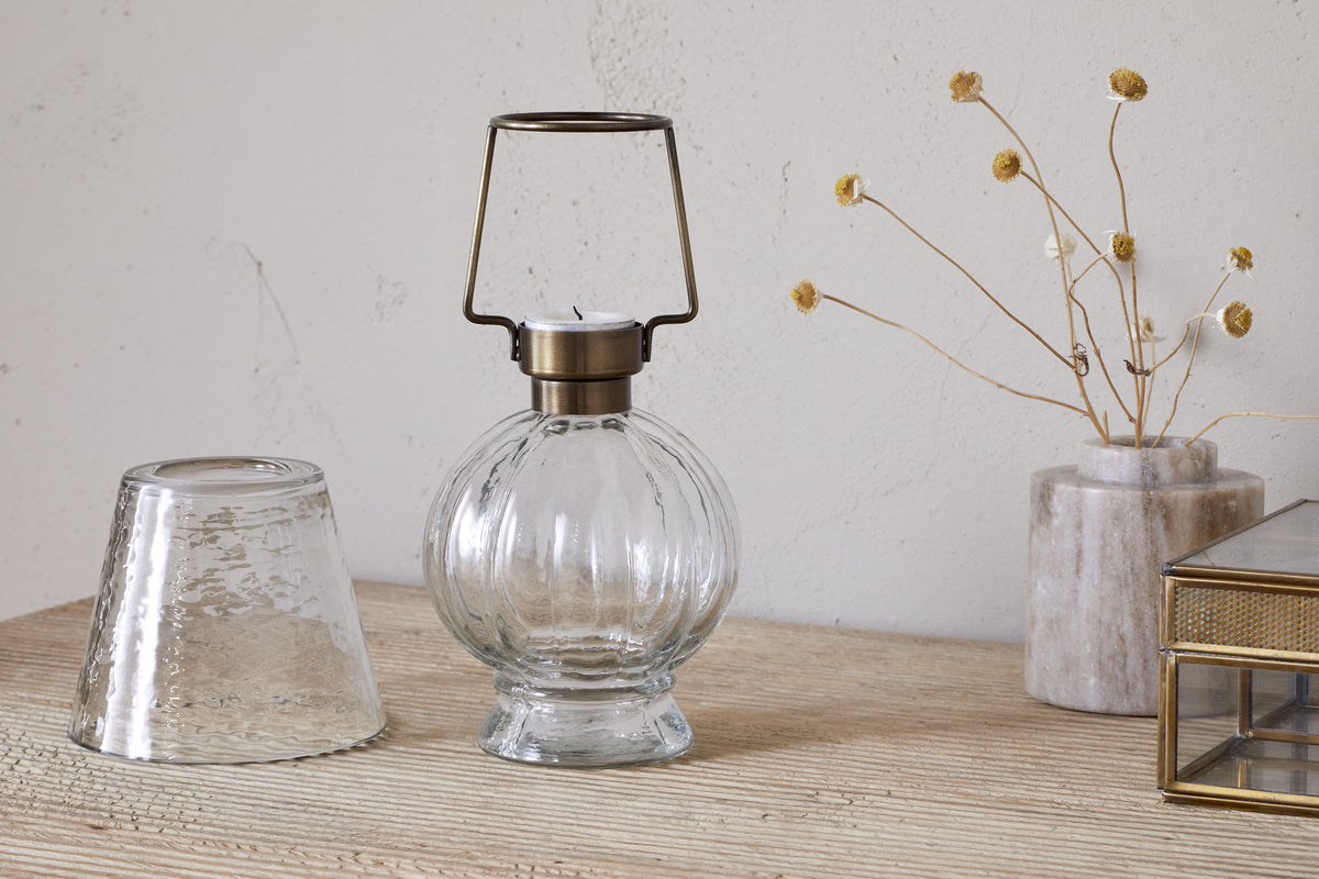 The nkuku Adesola Glass Tealight Holder Lamp in clear, featuring a brass handle, rests on wood alongside an upside-down glass, a marble vase with dried flowers, and a glass box before a textured light wall.