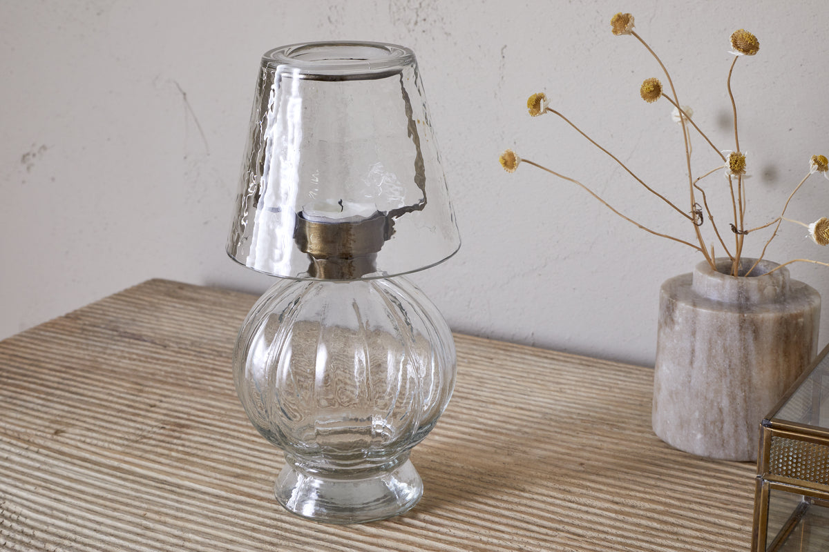 The nkuku Adesola Glass Tealight Holder Lamp in clear, featuring a round base and matching shade, is placed on a striped beige surface beside a vase of dried yellow flowers against a light wall.