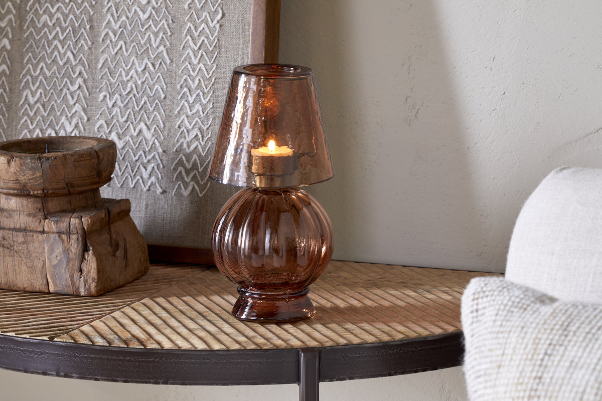The nkuku Adesola Glass Tealight Holder Lamp in amber sits on a wooden table near a textured artwork and rustic box, casting warm, non-electric light, with part of a pale sofa visible to the right.