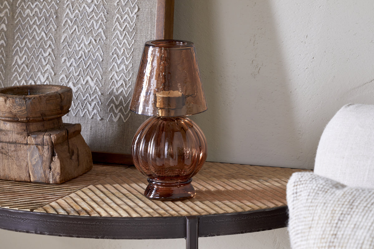 The Adesola Glass Tealight Holder Lamp - Amber by nkuku, with its rounded glass base and shade, sits on a wooden table beside rustic decor and a framed textile, near a beige wall and white chair—perfect for non-electrical lighting.