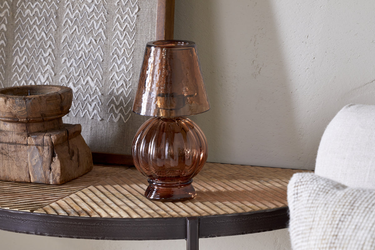 The nkuku Adesola Glass Tealight Holder Lamp in amber, featuring a ribbed base and matching shade, sits on a wooden table with rustic decor, tealight holders, and a woven textile in a cozy, neutral-toned room.