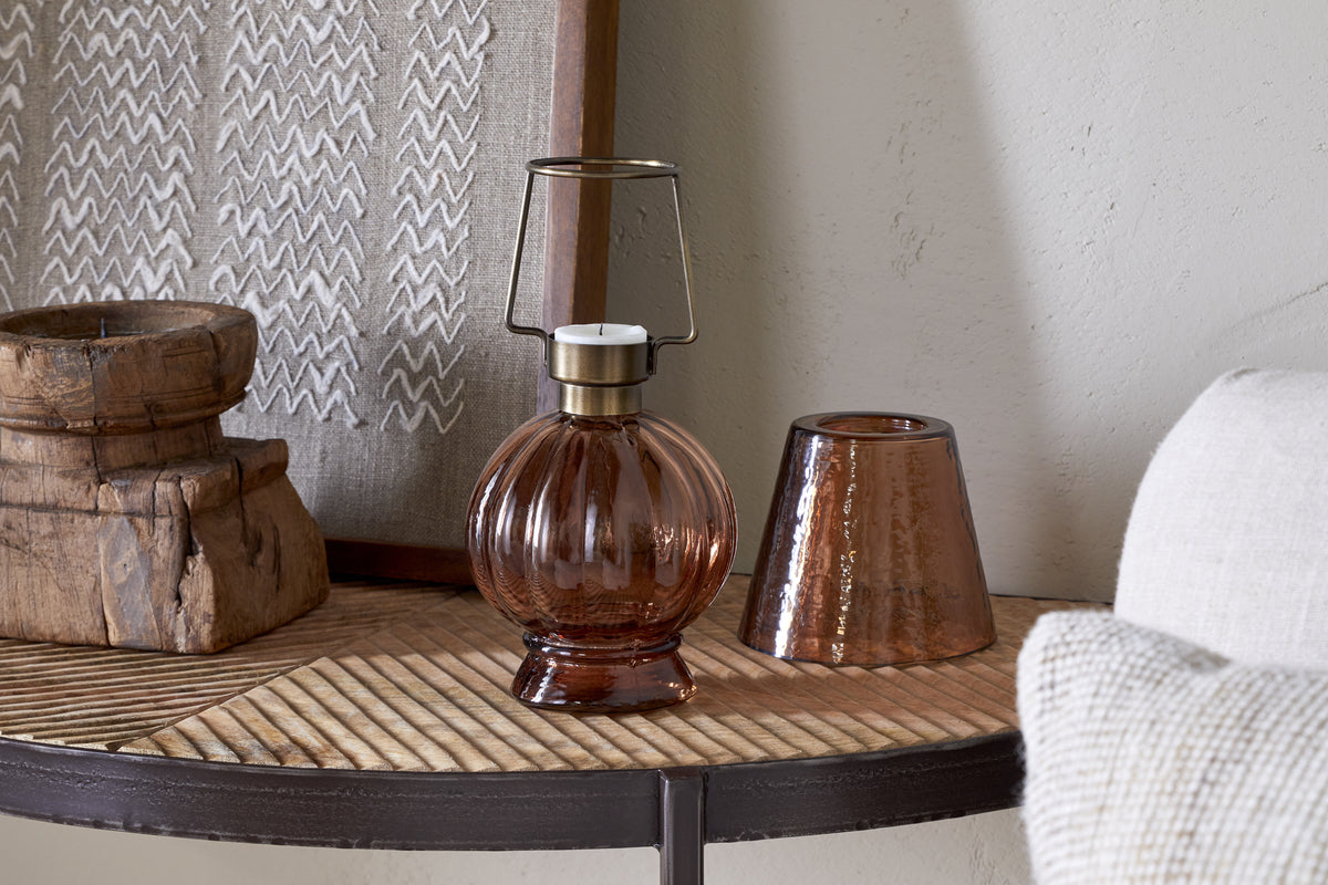The nkuku Adesola Glass Tealight Holder Lamp - Amber, with its round glass design and metal handle, sits on a wooden surface beside textured brown tealight holders and rustic decor—perfect for non-electrical lighting.