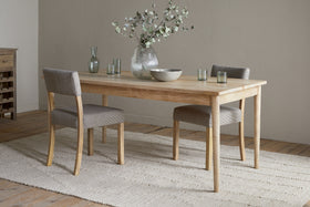 A minimalist dining room featuring the nkuku Anbu Mango Dining Table - Natural, two gray upholstered chairs, a textured rug, and a vase with green branches. Glassware and a bowl sit atop the elegant Anbu collection table.
