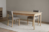 The Anbu Mango Dining Table - Natural by nkuku is paired with two gray upholstered chairs, placed on a beige textured rug in a minimalist space with wooden floors and light walls.