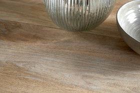 A close-up of the Anbu Mango Dining Table - Natural by nkuku, featuring a light wooden surface with a ribbed transparent glass vase and ceramic bowl partially visible in the top right corner.