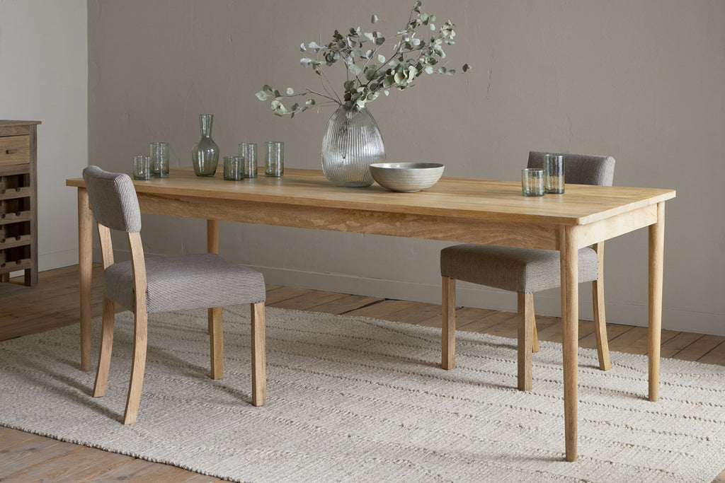 A minimalist dining room features the nkuku Anbu Mango Dining Table in natural finish, paired with two gray upholstered chairs, a large glass vase with branches, glassware, and a bowl on a textured rug over wooden flooring.