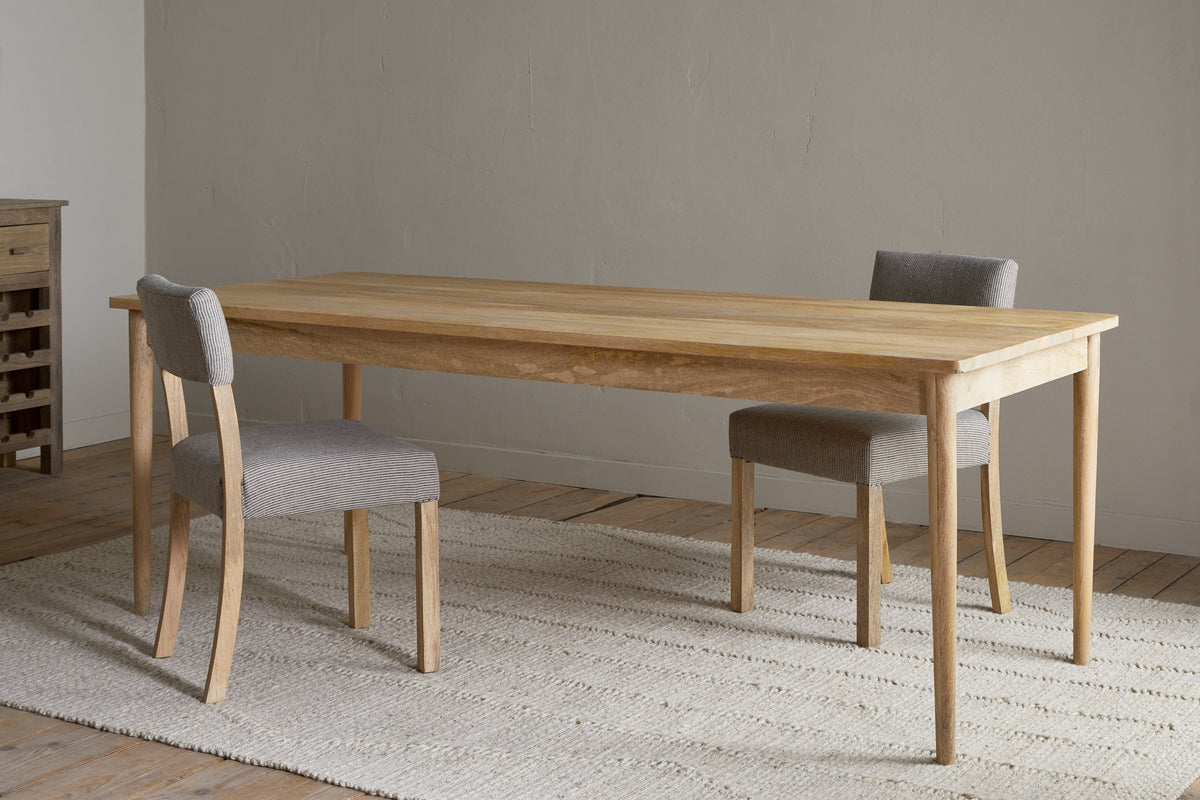 The Anbu Mango Dining Table - Natural by nkuku stands on a textured beige rug, paired with two gray upholstered chairs in a minimalist room featuring wooden floors and a plain light-colored wall.