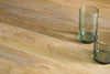 Two empty, transparent glass tumblers rest close together on the lower right corner of an nkuku Anbu Mango Dining Table in natural finish, showcasing the elegant wood grain and creating a refined dining atmosphere.