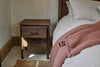 The nkuku Anbu Acacia Bedside Table in Washed Walnut, featuring two drawers, stands by the bed with striped bedding and a dusky pink blanket, offering extra storage and warmth to this softly lit, cozy bedroom.