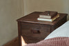 The nkuku Anbu Acacia Bedside Table in Washed Walnut features a closed drawer and displays two stacked books and a notebook with a pen, beside a bed with a brown knitted blanket, offering stylish bedroom storage.