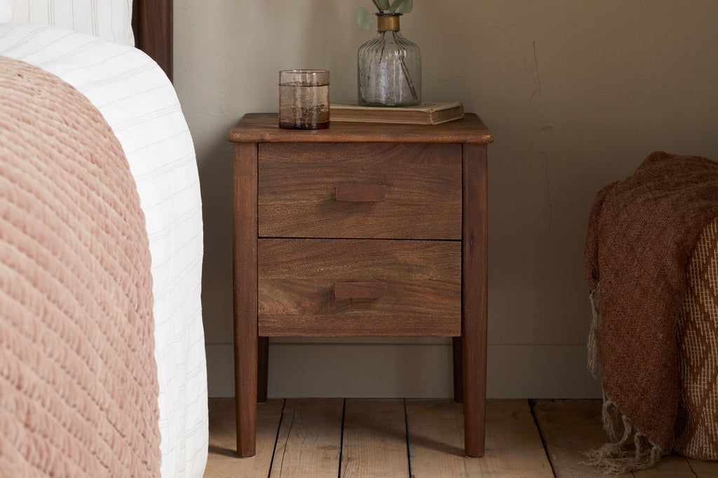 The nkuku Anbu Acacia Bedside Table - Washed Walnut stands by a bed with a textured pink blanket, topped with a glass, book, and a vase holding a single stem—adding stylish storage to the wooden-floored bedroom.
