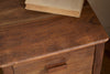 A close-up of the nkuku Anbu Acacia Bedside Table - Washed Walnut shows a book on top, smooth brown walnut finish, visible drawer handle, and stylish bedroom storage.