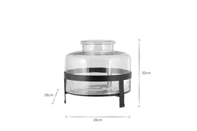 The Akoro Recycled Glass Vase - Clear by nkuku is a cylindrical glass jar (32cm high, 28cm wide) with a wide opening, displayed on a black three-legged metal stand that matches the width and elevates the vase by 28cm.