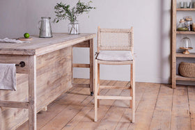 A rustic kitchen features nkuku's Aamani Mango Wood & Cord Counter Chair in natural by a wooden counter holding a pitcher, glass, vase with greenery, and lime. Shelves above display jars, baskets, and covered dishes.