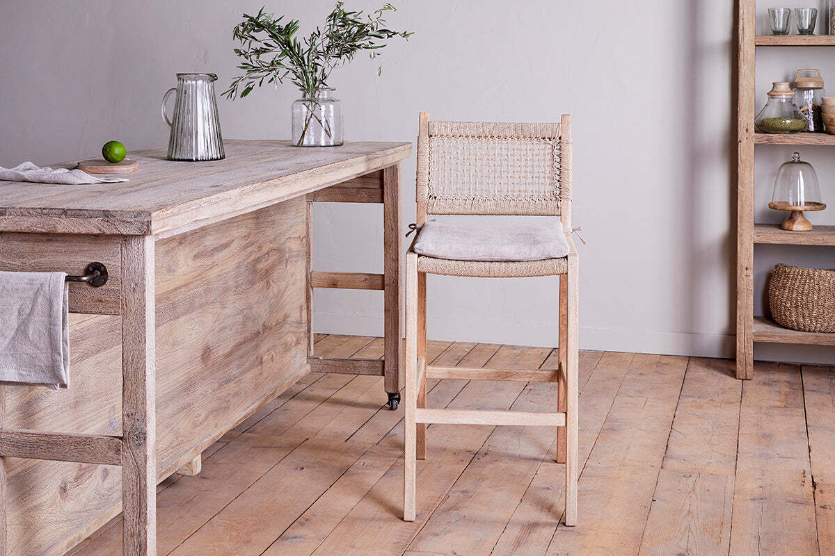 A rustic kitchen features nkuku's Aamani Mango Wood & Cord Counter Chair in natural by a wooden counter holding a pitcher, glass, vase with greenery, and lime. Shelves above display jars, baskets, and covered dishes.