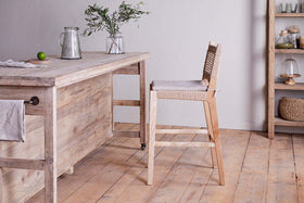 A rustic kitchen features nkuku's Aamani Mango Wood & Cord Counter Chair in natural, paired with a light wood table holding a glass jug, greenery-filled vase, and lime. Shelves display jars, bowls, and a glass dome above light wood flooring.