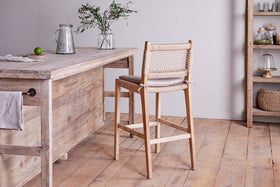 An Aamani Mango Wood & Cord Counter Chair by nkuku sits at a rustic kitchen island topped with a glass jug, lime, and a small vase of greenery. Shelves in the background display jars, baskets, and dishes for added charm.
