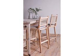 Two nkuku Aamani Mango Wood & Cord Counter Chairs in natural sit by a light wood counter topped with glassware, plates, and a vase of green branches. A wooden floor and plain light wall complete the serene setting.