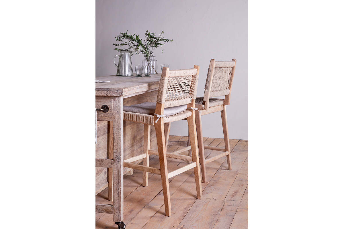 Two nkuku Aamani Mango Wood & Cord Counter Chairs in natural sit by a light wood counter topped with glassware, plates, and a vase of green branches. A wooden floor and plain light wall complete the serene setting.