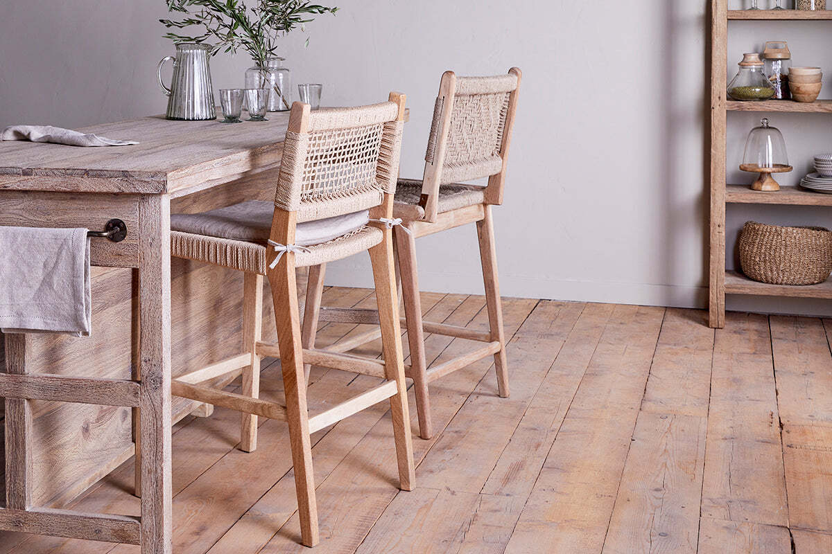 A light wood kitchen features nkuku's Aamani Mango Wood & Cord Counter Chairs—Natural, with woven backs at a rustic island. Clear glassware, a pitcher, and greenery accent the minimalist countertop and airy decor enhances the inviting space.