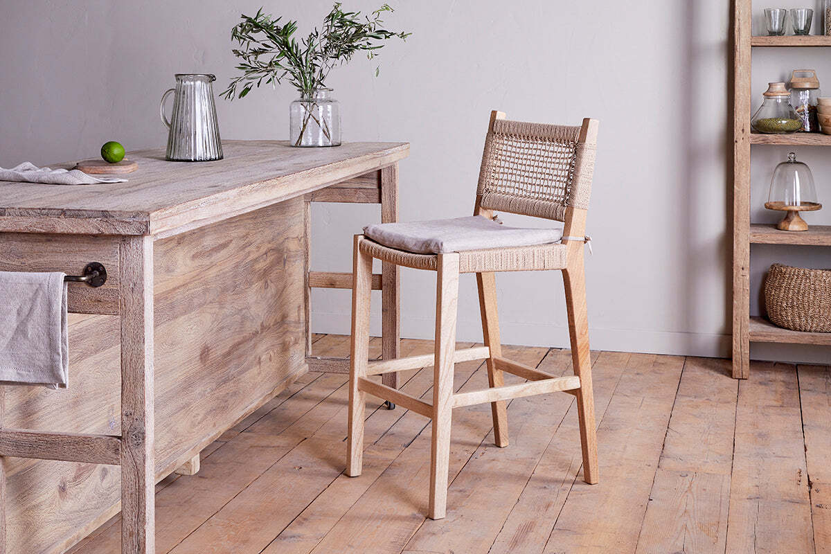 A rustic kitchen features the nkuku Aamani Mango Wood & Cord Counter Chair – Natural, showcasing a recycled paper cord woven back and cushioned seat beside a wooden island with a glass pitcher, vase of greenery, lime, shelving, and baskets in the background.