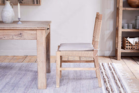 The Aamani Mango Wood & Cord Dining Chair by nkuku, crafted from natural mango wood with a beige cushion, pairs with a matching table. A woven rug, shelf of arranged dishes, and soft lighting complete the rustic, neutral-toned space.
