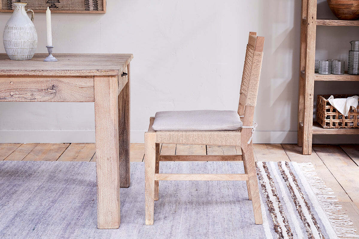 The Aamani Mango Wood & Cord Dining Chair by nkuku, crafted from natural mango wood with a beige cushion, pairs with a matching table. A woven rug, shelf of arranged dishes, and soft lighting complete the rustic, neutral-toned space.