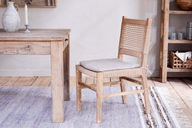 A light wood dining table with a carved pitcher & candle sits next to the nkuku Aamani Mango Wood & Cord Dining Chair - Natural, featuring a light grey cushion, on a striped rug. A rustic wooden shelving unit is on the right.