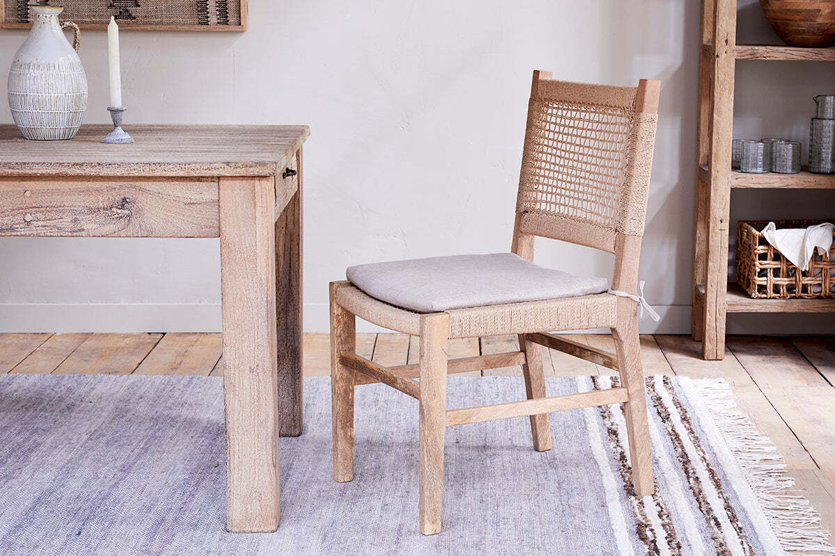 A light wood dining table with a carved pitcher & candle sits next to the nkuku Aamani Mango Wood & Cord Dining Chair - Natural, featuring a light grey cushion, on a striped rug. A rustic wooden shelving unit is on the right.