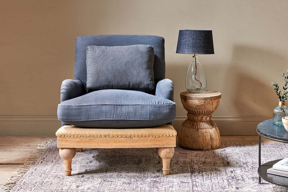 A cozy living room corner features the nkuku Abe Deconstructed Linen Armchair in charcoal, accented by a cushion, a mango wood ottoman, a rustic round wooden side table with glass lamp and dark shade, part of a round marble table, patterned rug, and beige walls.