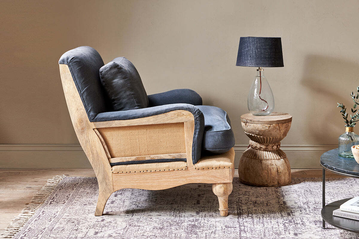 A side view of the nkuku Abe Deconstructed Linen Armchair in charcoal, featuring a mango wood frame, sits beside a carved side table with a clear glass lamp and dark shade on a patterned rug, with a coffee table and plant nearby.