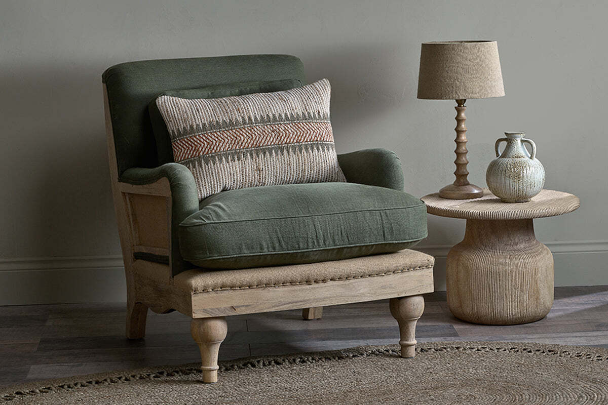 An Abe Deconstructed Linen Armchair - Olive by nkuku, with green cushions, sits beside a round wooden side table holding a beige lamp and ceramic jug. The minimalist space features earthy tones and a woven rug.