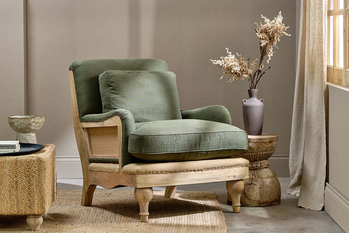 The Abe Deconstructed Linen Armchair in olive by nkuku features plush cushions and light wooden legs. Placed beside a rustic side table with a dark vase of dried flowers, it glows in sunlight for a warm, inviting atmosphere.