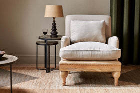 The Abe Deconstructed Linen Armchair - Stone by nkuku sits atop a woven rug. Next to it, a round black side table holds a tan-shaded lamp and glass of red wine, all set against green curtains for an inviting atmosphere.