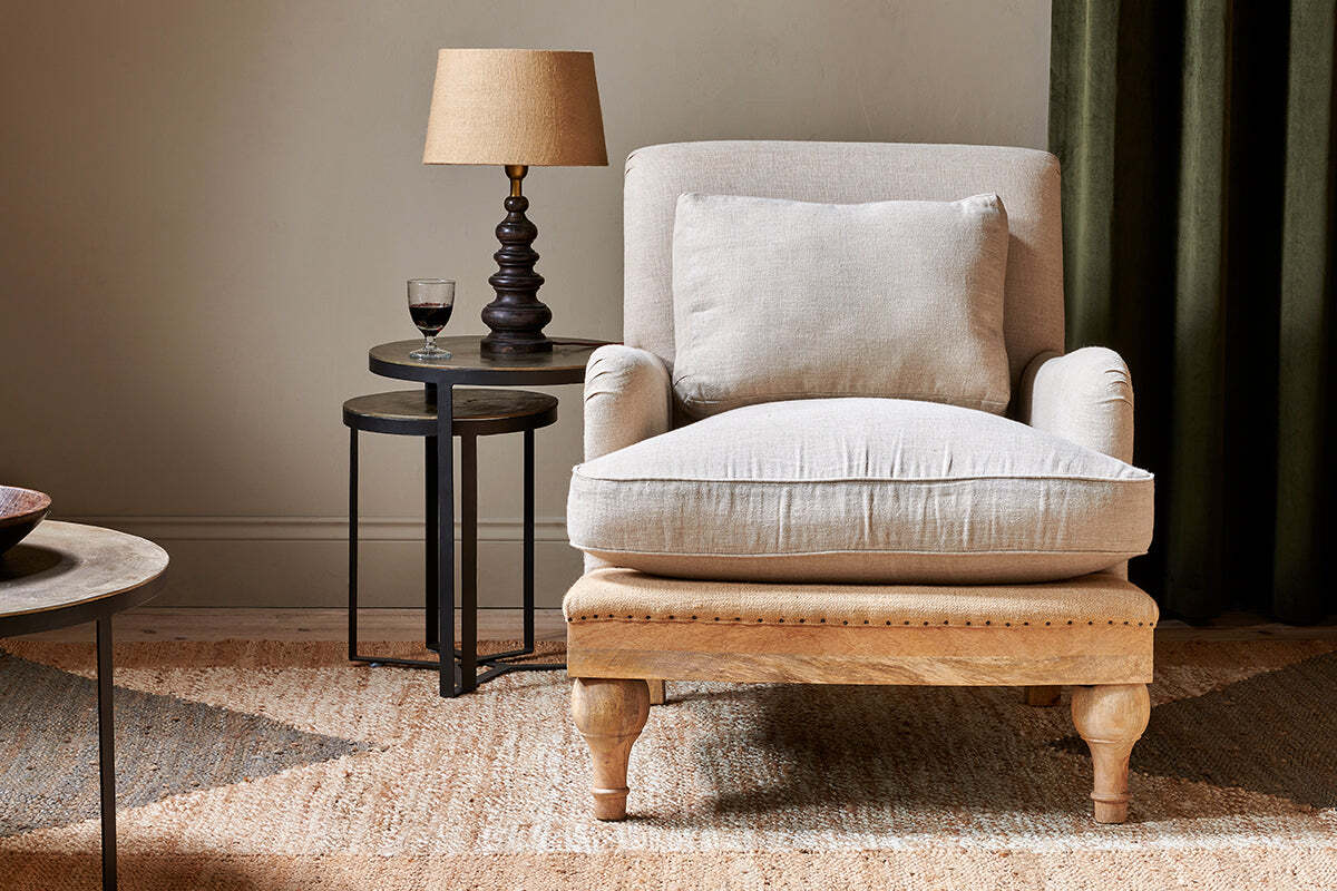 The Abe Deconstructed Linen Armchair - Stone by nkuku sits atop a woven rug. Next to it, a round black side table holds a tan-shaded lamp and glass of red wine, all set against green curtains for an inviting atmosphere.